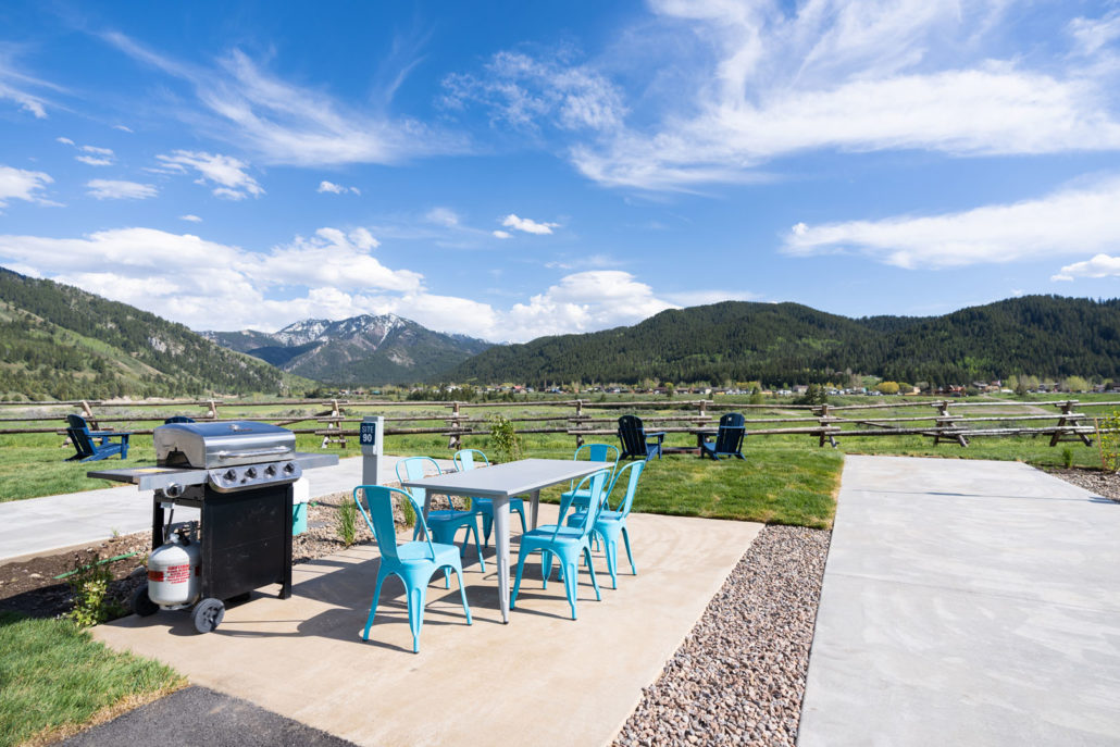 A premium RV campsite with a grill, blue dining chairs, a white dining table, and mountains.