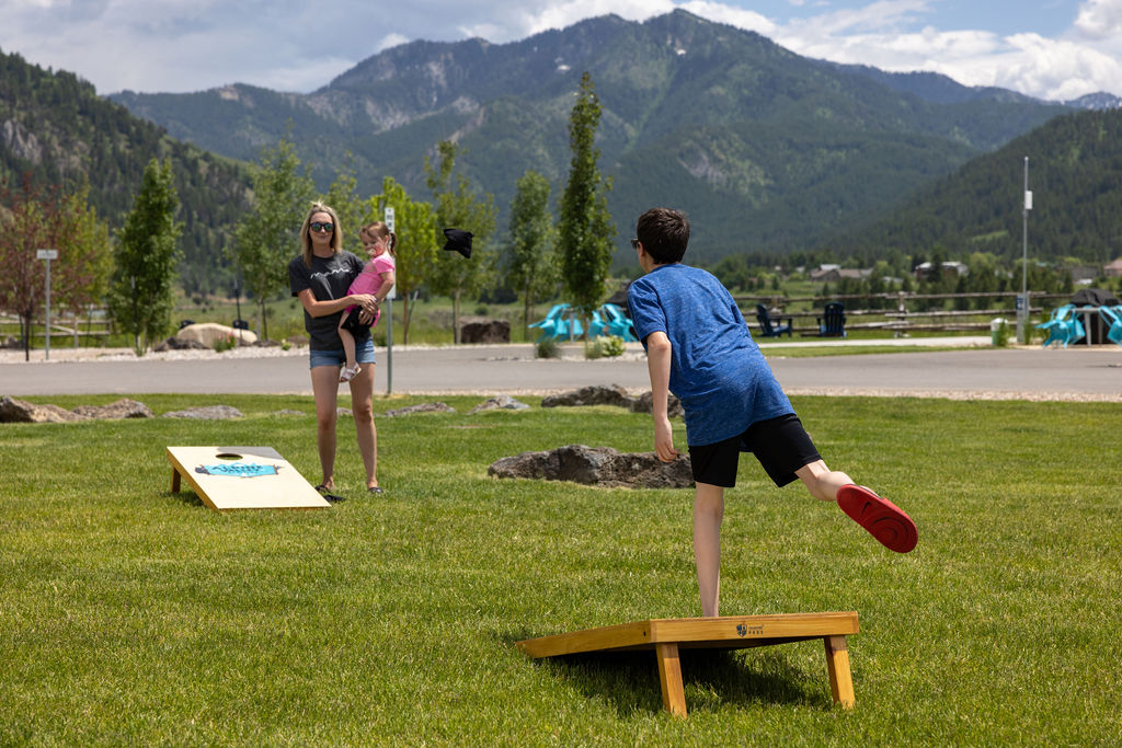 A family of three plays corn hole on a green lawn in front of large mountains.