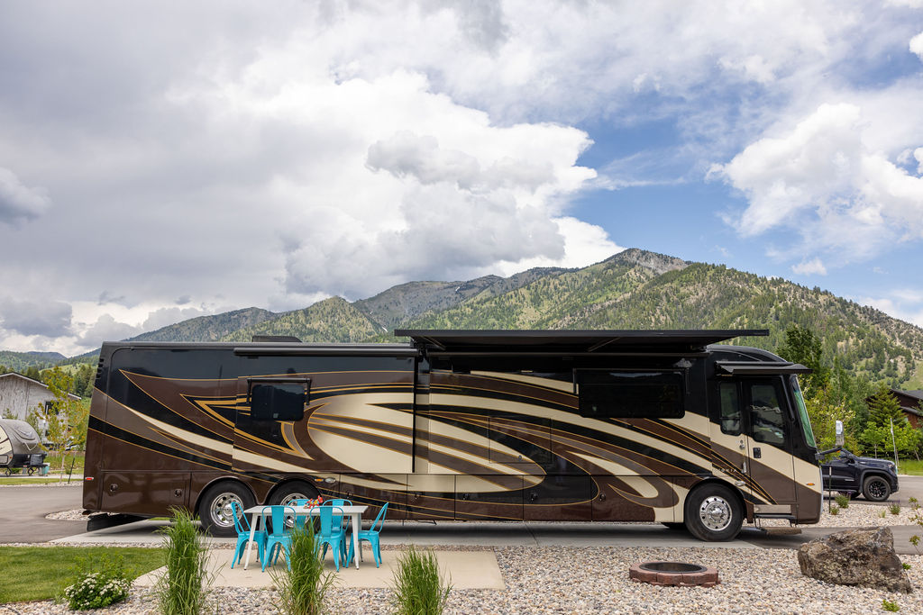 A large, brown RV parked in a pull-through campsite with mountains in the background.