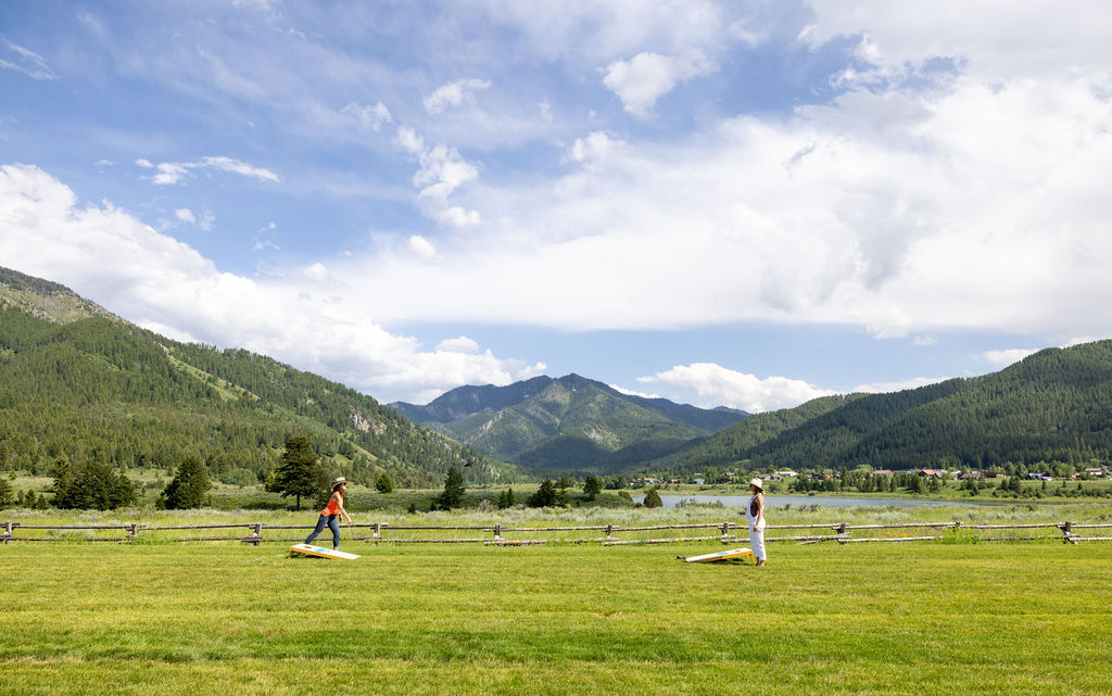 Two women playing a yard game on green grass with mountains in the background.